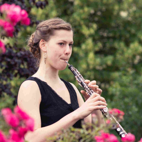 Die Oboistin Elisabeth Beckert spielt in einem scharzen Kleid hinter einer Blumenhecke ihr Instrument